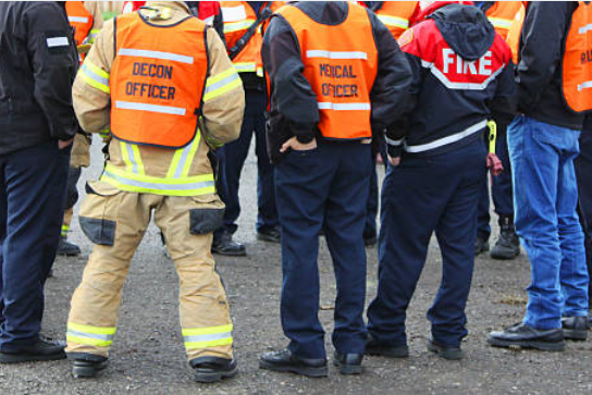 Photograph of the backs of a group of first responders including some wearing orange safety vests imprinted with text naming their response roles - decon officer, medical officer.