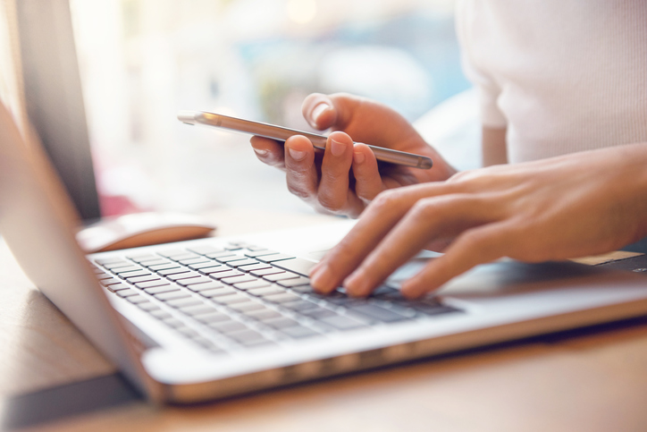 Photographic close-up image of a person's left hand on a laptop keyboard and their right hand holding a mobile phone.